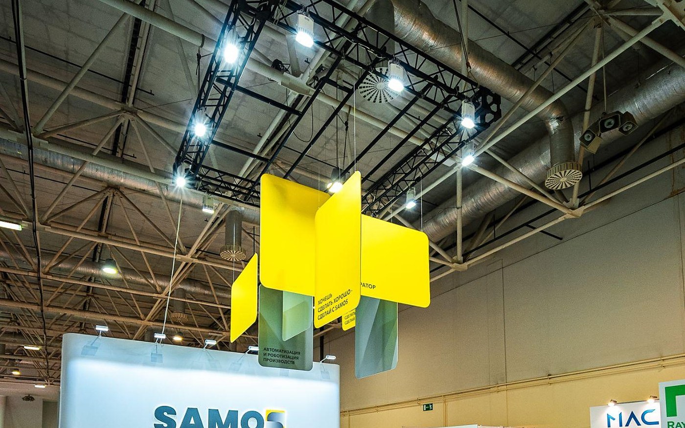 Подвесная жёлтая конструкция со знаком SAMOS над выставочным стендом в павильоне